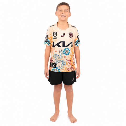 A young boy stands barefoot against a white background wearing a short-sleeve patterned rugby jersey and black shorts. The jersey has a cream base with colorful abstract and floral designs, along with visible ASICS branding.