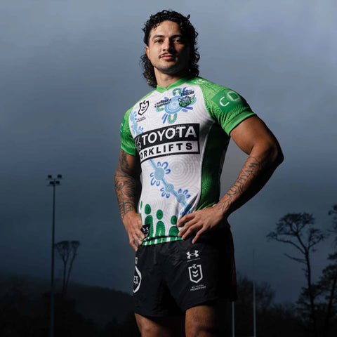 A male rugby player wearing a green and white Canberra Raiders indogenous jersey with black shorts, posing outdoors against a dark, moody sky.