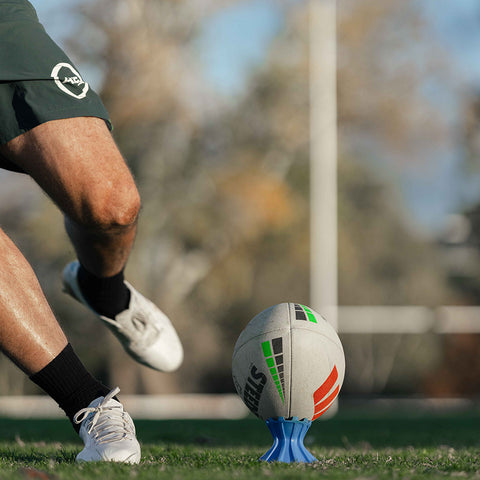 A close-up action shot of a rugby ball on a blue stand on a grassy field, with a player's legs in dark green shorts and white cleats blurred in the background.