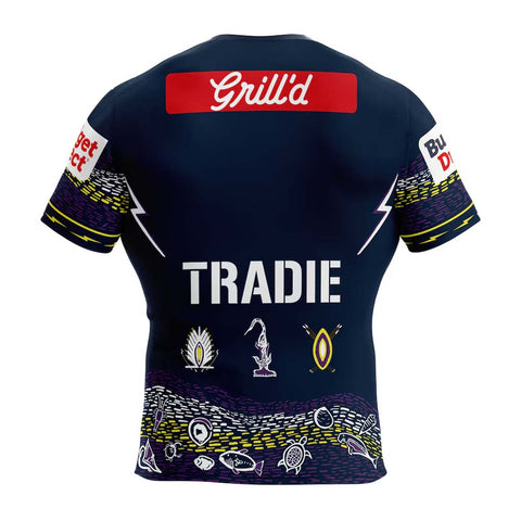 Back view of a navy blue short-sleeve athletic jersey with 'TRADIE' printed in large white letters across the center. The jersey features a red sponsor logo 'Grill'd' on the upper back, white text on the sleeves, and a detailed Aboriginal-inspired wave pattern in purple, yellow, and white along the bottom hem.