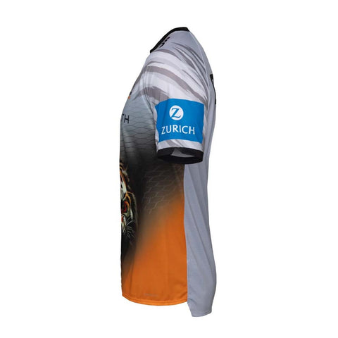 A wests tigers 2026  coaches tee shown from the side profile on a white background, featuring a gray and black patterned upper section, a blue sleeve patch with the white 'ZURICH' logo, and an orange gradient lower section with a tiger graphic.